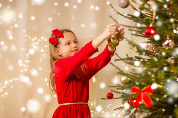 christmas, holidays and childhood concept - happy girl in red dress decorating natural fir tree