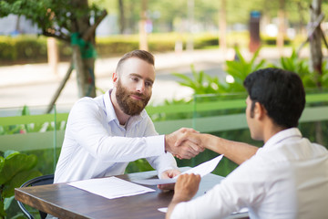 Confident man making handshake with new business partner in outdoor cafe. Handsome bearded businessman in white shirt satisfied with new collaboration. Concluding deal concept