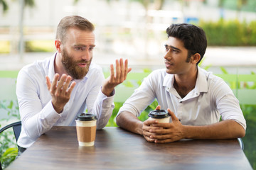 Two male friends drinking coffee and talking in outdoor cafe. People sitting at table with blurred...