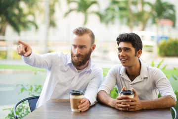 Two male friends drinking coffee in outdoor cafe. One man showing something away with blurred plants in background. Coffee break concept. Front view.
