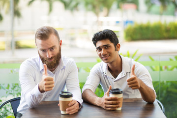 Two men drinking coffee and showing thumbs up in outdoor cafe. People looking at camera with blurred plants in background. Coffee break concept. Front view.