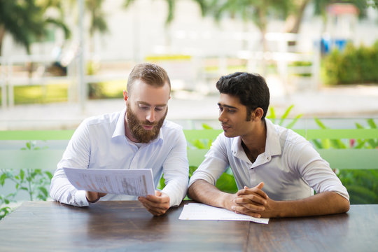 Two focused business men meeting and working in outdoor cafe. Multiethnic businesspeople sitting at table and discussing diagrams with blurred plants in background. Teamwork concept. Front view.