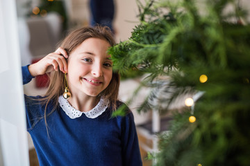 Small girl standing by a Christmas tree at home, a ball ornament on her ear. Copy space.