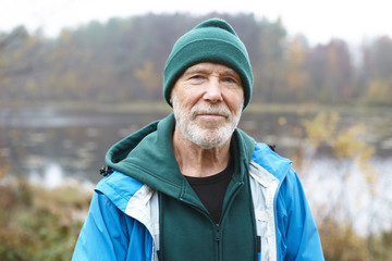 Naklejka na ściany i meble Outdoor close up shot of handsome active elderly male pensioner with gray beard having morning walk in wild nature, posing against misty lake and colorful autumn forest background, looking at camera