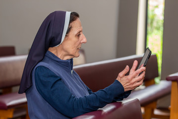 Religious Nun kneeling  in a chapel pew while looking at a tablet.