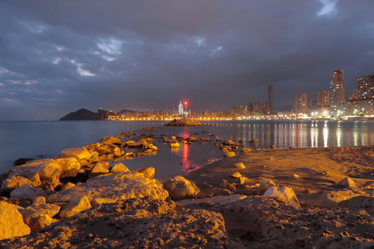 Night landscape on Poniente beach with rocks, lighthouse and buildings lights reflected in the water. Nightfall in Benidorm, Spain, with cloudy sky