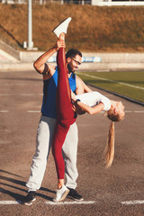 Multicultural handsome bearded man in blue shirt help stretching to his blonde athletic girlfriend at the football stadium outdoor, healthy lifestyle and people concept