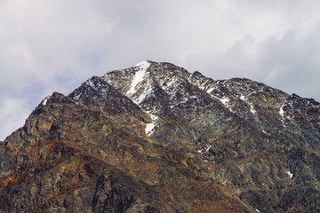 Snowy mountain top in cloudy sky. Rocky ridge under clouds. Overcast weather in highlands. Atmospheric minimalist landscape of majestic nature.
