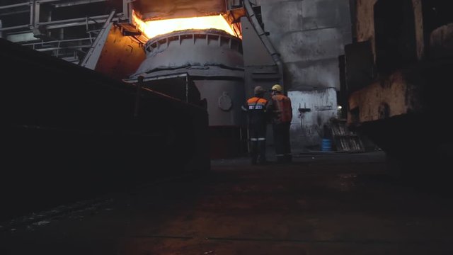 View of two workers from back in uniform and helmets on background of dark dirty factory. Footage. Two employees monitor and control work of production