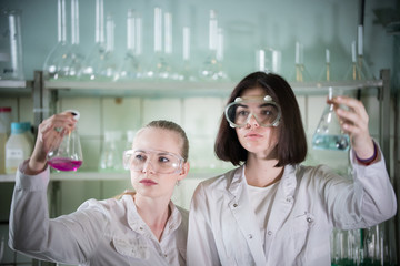 Chemical laboratory. Two young woman holding different flasks with liquids in it. Looking at the flask