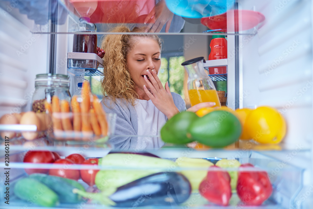 Wall mural Young Caucasian woman yawning and taking juice from fridge. Morning routine. Picture taken from the inside of fridge.