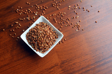 White Bowl with grains of buckwheat on a wooden table, top view. Randomly scattered grains around the bowl. 