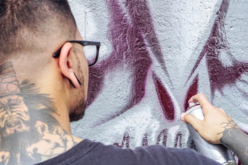 Street graffiti artist painting with a color spray can a dark monster skull graffiti on the wall in...