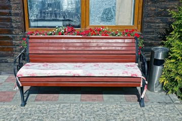 brown wooden bench stands on the sidewalk near the wall of the house with a window and flowerpots with flowers