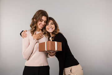 Image of two cheerful girls smiling and posing with present box, isolated over gray background