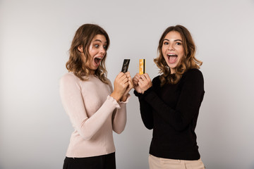 Image of two beautiful women smiling and holding credit cards, isolated over gray background