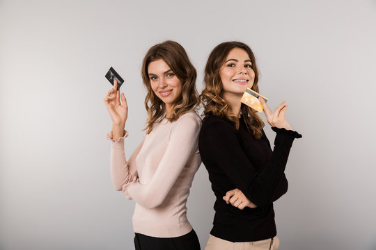 Image of two happy women standing back to back and holding credit cards, isolated over gray background
