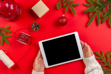 Christmas composition. Top view of tablet computer in woman hands. Christmas decoration and green fir branches on red background