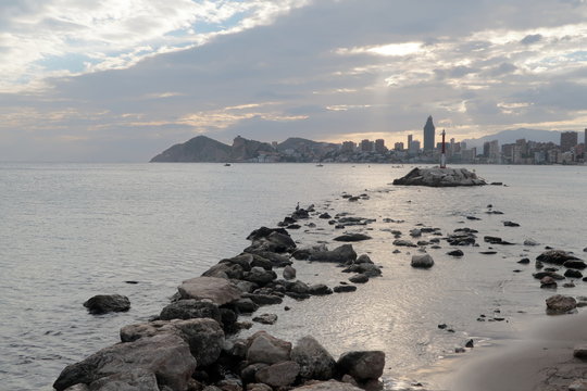 Meditarranean sea landscape in Benidorm at sunset. Rocks and the lighthouse on a small island with buildings in the background on a cloudy day.