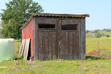 Old dilapidated wooden garage with two doors and missing door handle now used as garden shed for storing tools surrounded with uncut grass mixed with small flowers and large tree with family houses in