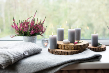 Fototapeta na wymiar Ready for autumn. Common heather flower in zinc pot, home decor idea. Set of cozy seasonal decorations on window sill. Gray candles lit, wooden boards, common pink heather flower in flower pot.