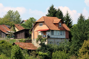 Fototapeta na wymiar Unfinished red brick suburban family house located on top of the small hill next to wooden garden tool shed surrounded with other older houses, tall trees and other forest vegetation with cloudy blue 