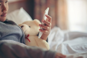 Asian child sitting and using smart phone via 4g internet to watching movies in bedroom.Relationship of Cute little girl hugging teddy bear and playing game with smartphone on a white bed at home.