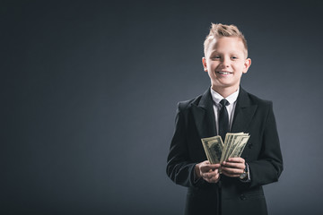 portrait of smiling preteen boy dressed as businessman holding dollar banknotes in hands on grey...