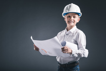 portrait of smiling boy in architect helmet holding blueprints on dark background