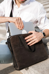 Photo of european man 30s wearing sunglasses, putting laptop into his bag, while sitting on stairs on city street