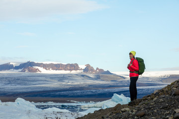 Fototapeta premium Young sporty girl with backpack admires beautiful view of the ice lake and mountains. Ice lagoon in Iceland