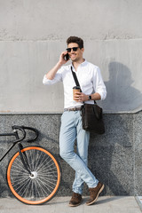 Photo of happy man 30s wearing sunglasses, drinking takeaway coffee and talking on mobile phone while standing with bicycle along wall outdoor