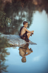 Caveman boy sitting on the rock near river or lake and looking away. Evolution survival concept. Creative art fantasy photo