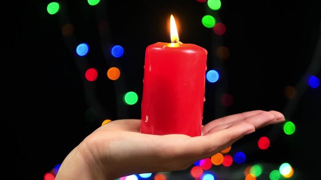 Close-up of female hand with a big red candle on bokeh background. Burning candle on a black background. Religion, spirituality and celebration.
