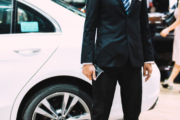 Fototapeta na wymiar An unidentified man in a black vest holding a notebook was walking across a black modern car. Handsome elegant man in suit, businessman or salesman, stretches his hand to conclude a business.