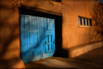 Naklejka premium Bright blue wooden doors and Adobe walls of this Santa Fe, New Mexico street view with shadows of trees and natural light of this high altitude plateau.