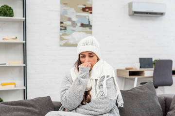 freezed young woman in warm clothes with runny nose sitting on couch at home
