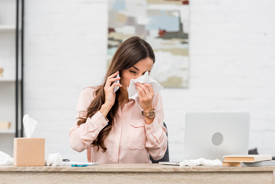 sick young businesswoman talking by phone and sneezing at workplace