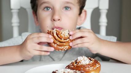 Happy boy is sitting behind the table and eating a cinnamon bun - Powered by Adobe