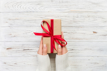Woman hands give wrapped valentine or other holiday handmade present in paper with red ribbon. Present box, decoration of gift on white wooden table, top view with copy space