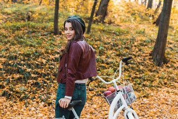 Fototapeta na wymiar selective focus of fashionable female model in beret and leather jacket posing near bicycle in autumnal forest