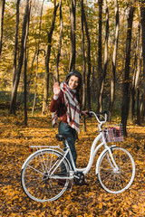 Fototapeta na wymiar smiling stylish girl in leather jacket and beret waving by hand and carrying bicycle in autumnal park