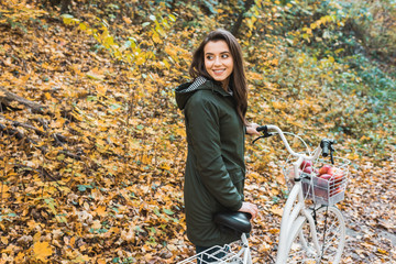 Fototapeta na wymiar smiling young woman carrying bicycle with basket full of apples in yellow autumnal forest