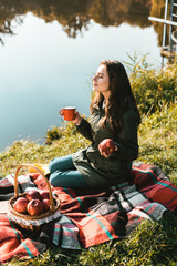 Cheerful girl with apple and cup of coffee sitting on blanket near pond