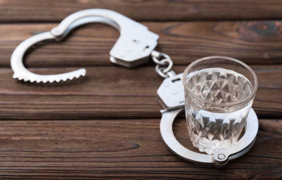 A glass of vodka, iron handcuffs to detain criminals on the background of a wooden table. Driving under the influence, crime of driving a car. offence.