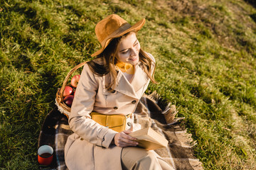 high angle view of smiling elegant woman in hat sitting on blanket and reading book outdoors