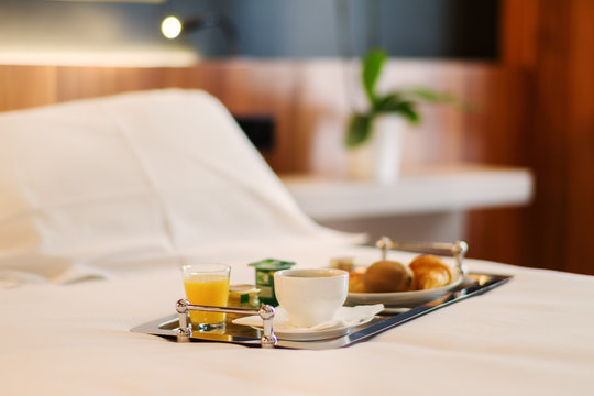 Plateau de petit-d&eacute;jeuner contenant du caf&eacute;, jus d'orange, croissant et yaourt pos&eacute; sur le lit d'une chambre d'h&ocirc;tel recouvert d'un drap blanc avec une plante verte en arri&egrave;re plan