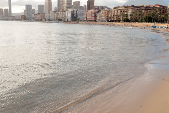Shore of the beach, calm crystal clear water with rays of the sun on the sand of Poniente beach overlooking the promenade and buildings