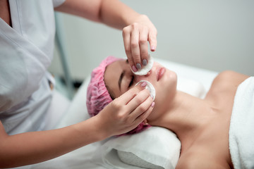 Young girl in white medical coach in cosmetological salon getting her face cleaned by proffecional cosmetologyst