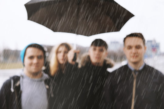 defocused group of young urban teenage friends under one umbrella in heavy rain - blurred background image with copy space - bad weather concept